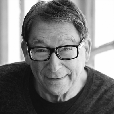 Black-and-white portrait of Stuart Weitzman wearing glasses and a dark sweater. He is smiling. 
